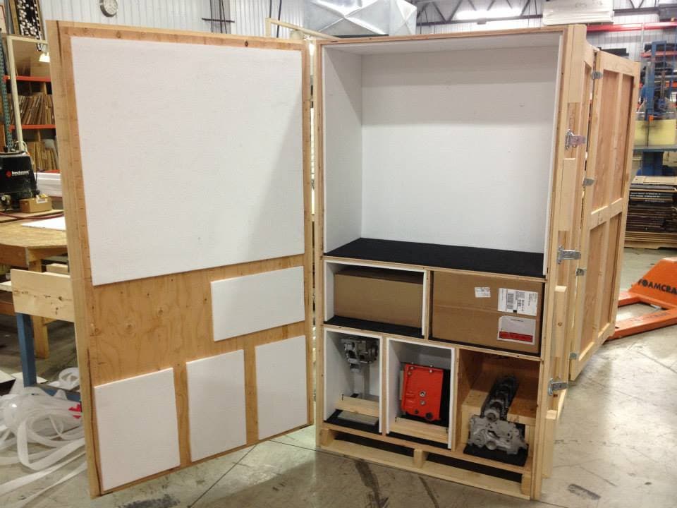 A large custom wood crate with foam-lined panels and internal compartments holding machined parts